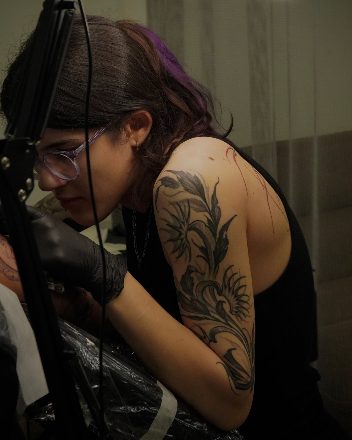 A tattoo artist wearing black gloves and glasses, focused on tattooing a client's upper arm with a floral design. The artist has dark hair with a purple streak and wears a black tank top.